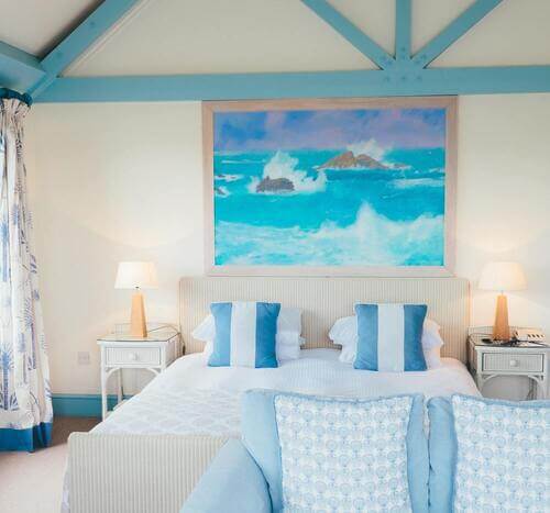 Chambre blanche avec moquette beige au sol et charpente en métal peinte en bleu au plafond. Grande hauteur sous plafond. Lit double avec lampe de chevet de part et d'autre. Fenêtres avec rideaux blancs et bleus. Grande peinture représentant la mer déchainée au dessus de la tête de lit.